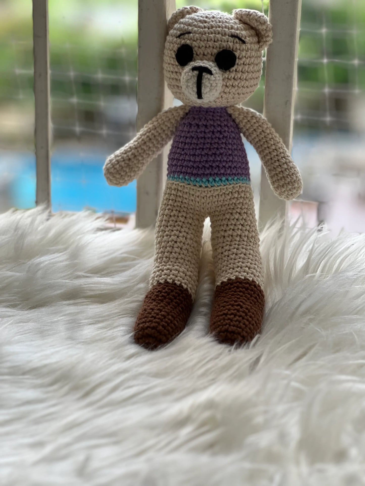 Handmade crochet teddy bear standing on white fluffy rug with outdoor blurred background, showing full body in purple striped top