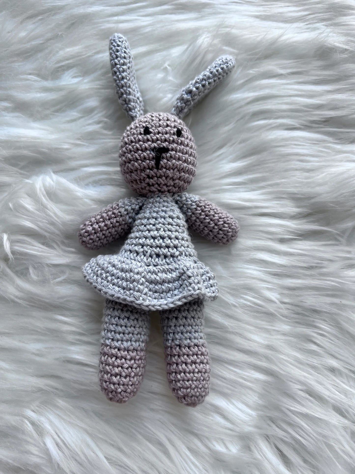 Gray handmade crochet bunny rabbit toy with dress standing upright on a soft white fluffy surface