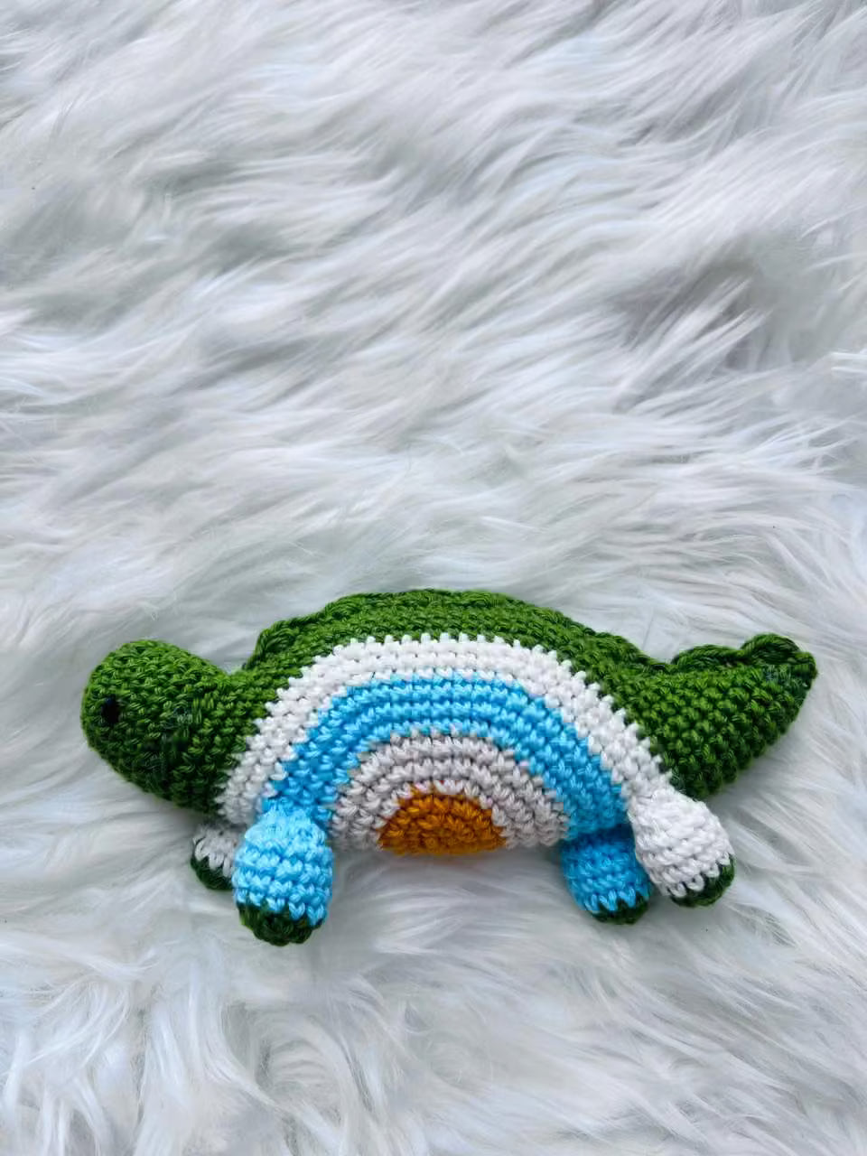 Handmade green crochet dinosaur toy with rainbow belly pattern on a fluffy white surface