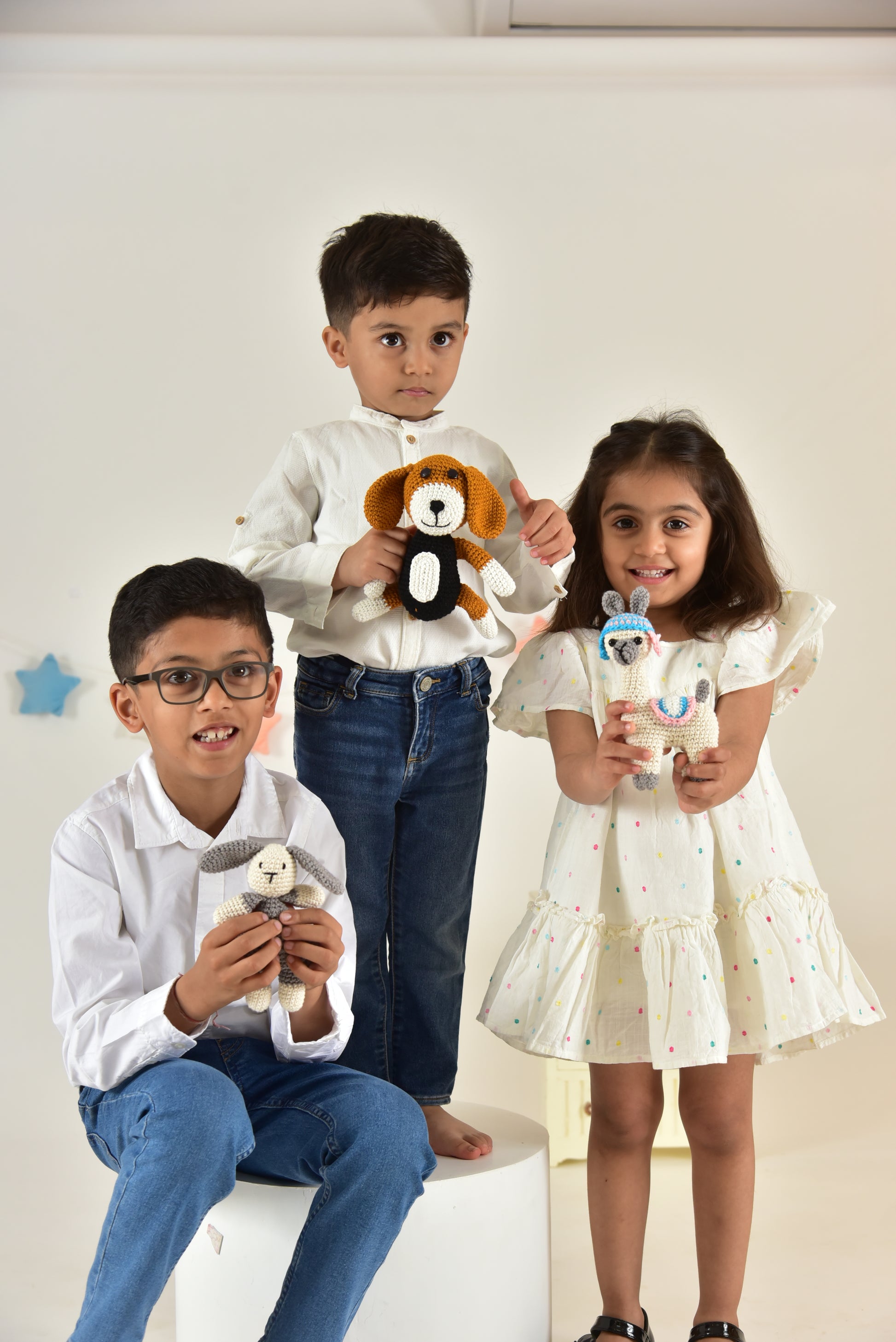 Three children in matching outfits displaying handmade crochet bunny and animal stuffed toys