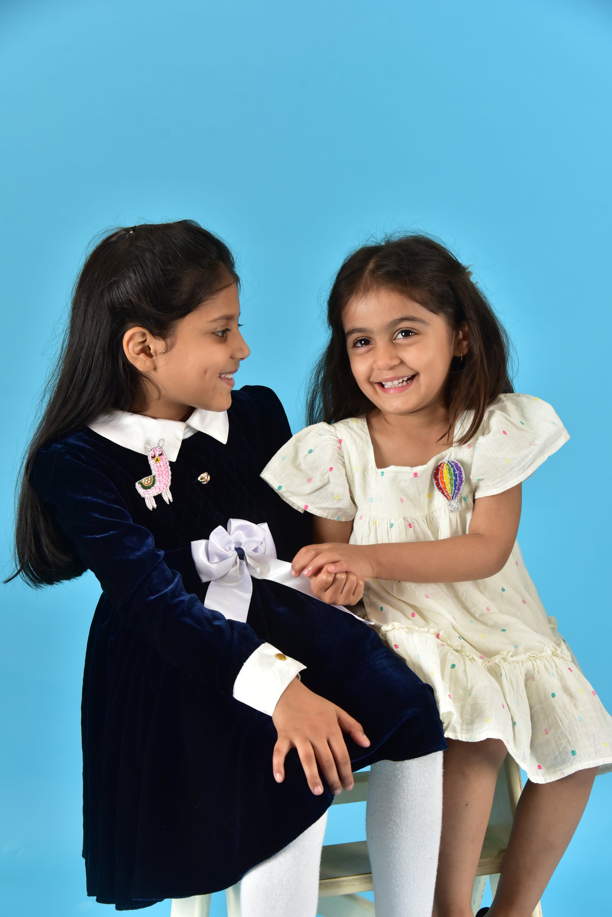 Two girls posing together against blue background with rainbow hot air balloon brooch worn on white dress