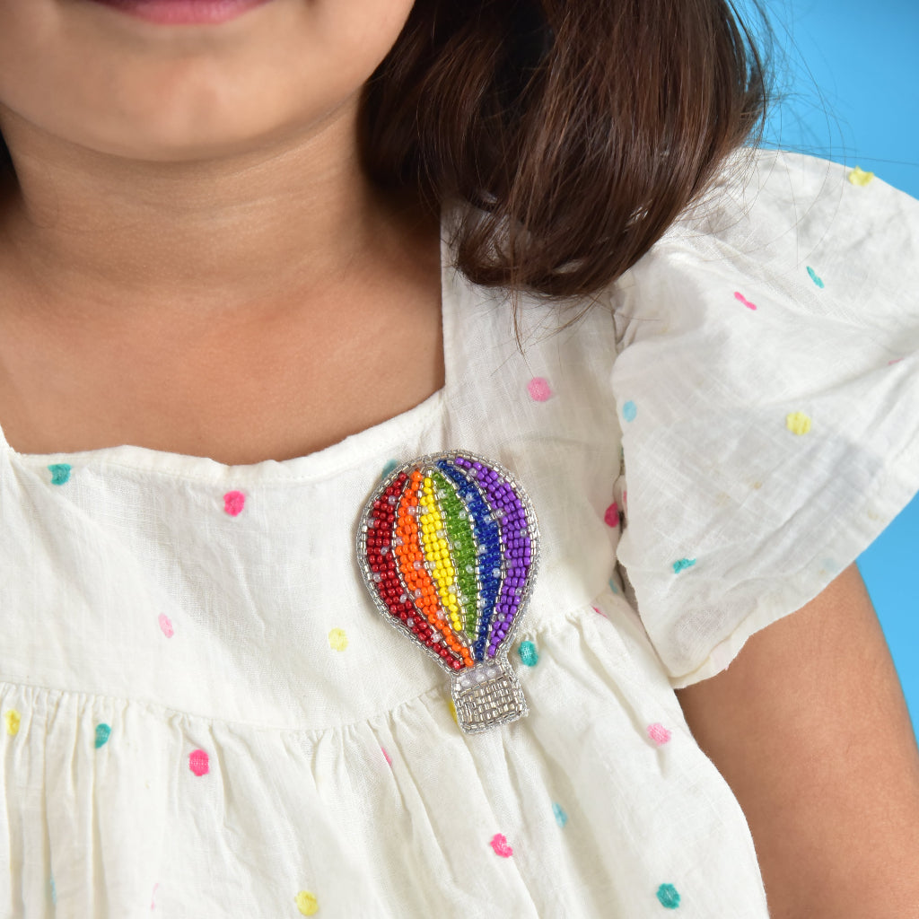 Child wearing rainbow hot air balloon brooch on white polka-dot dress against blue background