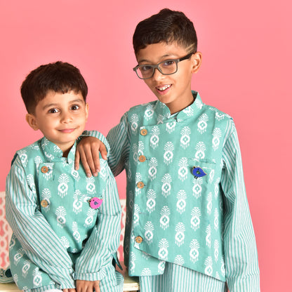 Two children wearing handcrafted bird brooches on matching teal traditional outfits against a pink background - Little Pink Llama
