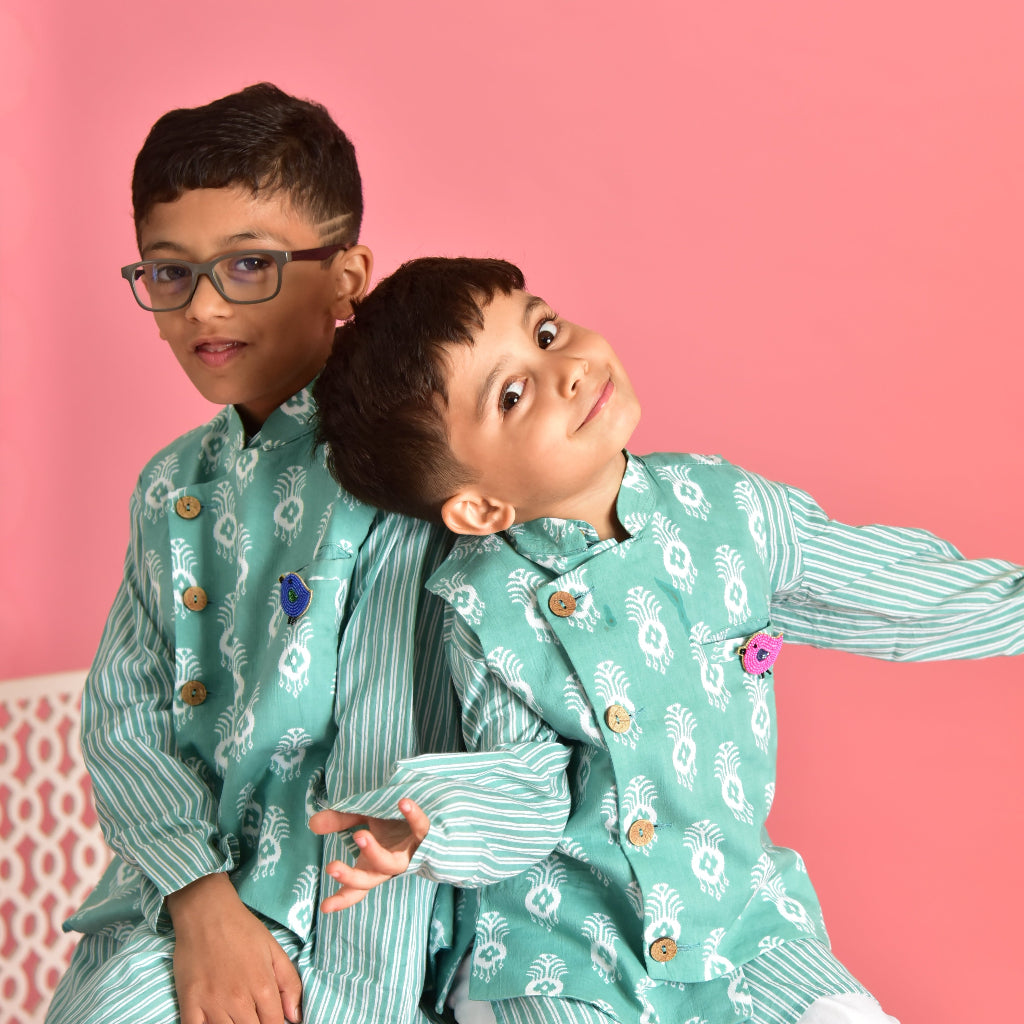 Two children wearing colorful bird brooches on matching teal traditional outfits against pink background - Little Pink Llama