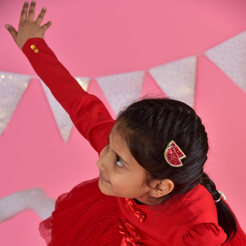Young girl in red dress wearing a handmade watermelon hair clip, arm raised, on pink festive background