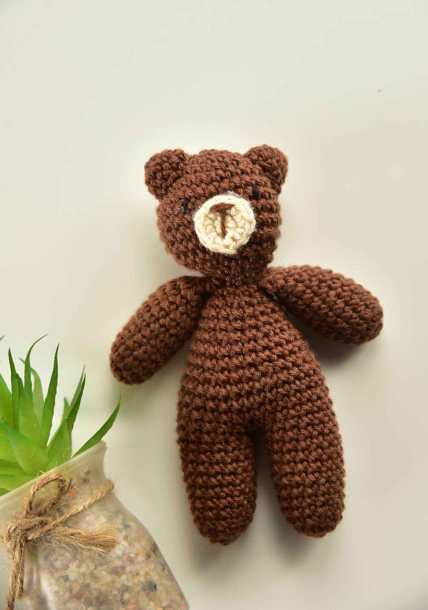 Mini brown crochet teddy bear displayed on light background with green plant - handmade cotton stuffed animal